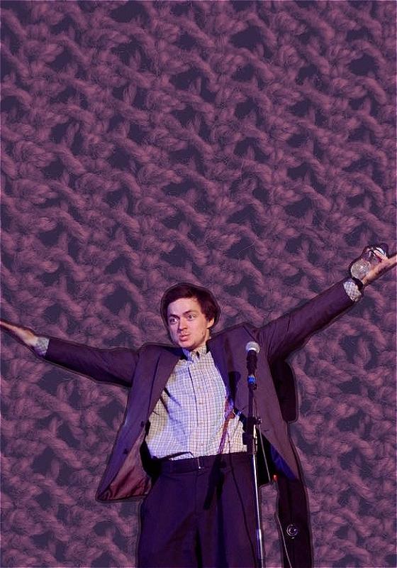 Tom is standing in front of a microphone, holding his arms out above him. He is wearing a check shirt and a purple suit jacket. The background is enlarged knit cloth in purple thread. 