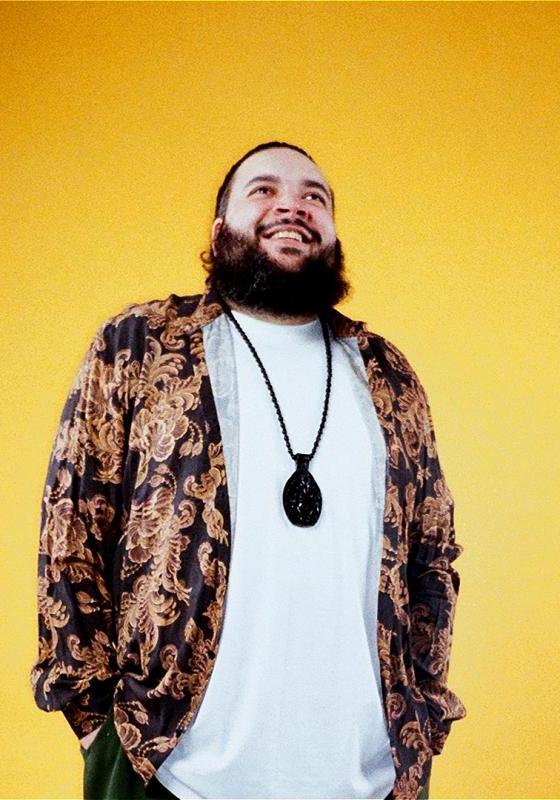 Raj wearing a white t shirt, with a black and gold patterned shirt on top, and an oversized necklace, stands against a bright orange backdrop, looking up and smiling. 