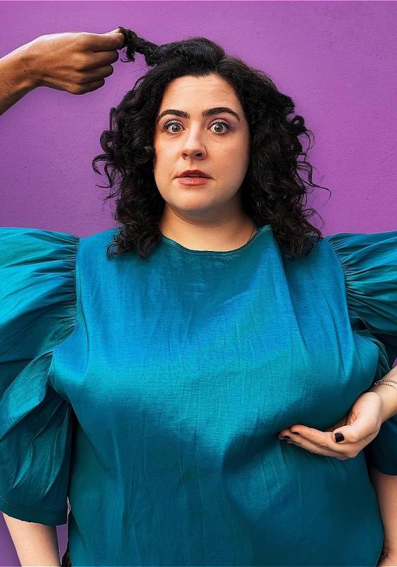 Jess Fuchs stands against a purple background, wearing a voluminous blue dress. Someone reaches into frame from the right and cups her breast. Someone else reaches in from the right and is holding a section of her black curly hair. 