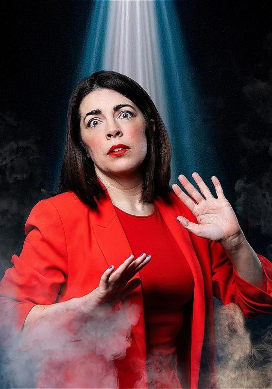 Rosie is wearing a red top and suit jacket, looking forward in fear. She is holding her hands up in defence and is surrounded by smoke. A beam of light shines down from above.