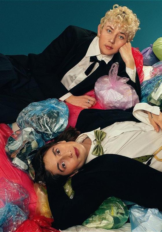 Two people in tuxedos are lying on top of a pile multicoloured bin bags