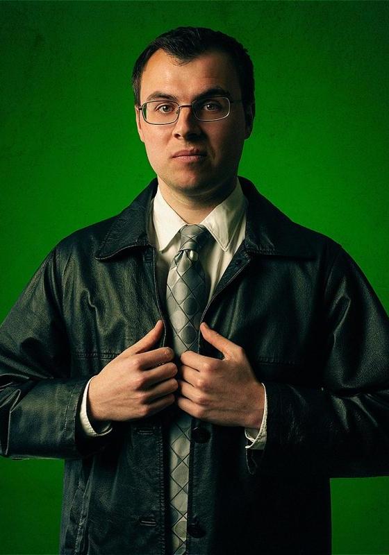 Freddie stands against a green backdrop looking straight ahead. He is wearing a shirt, tie, and a leather jacket. 