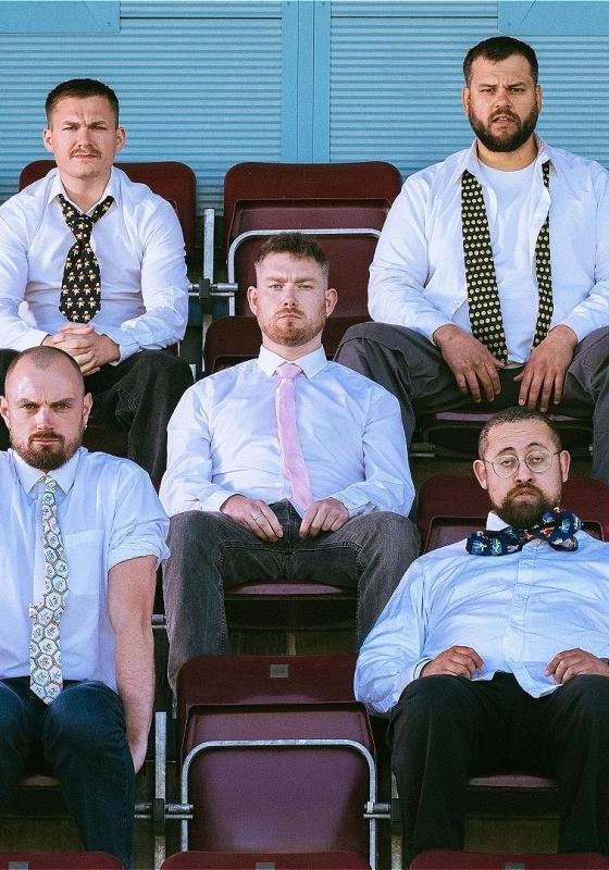Five men sit in bleacher style seats, all wearing a shirt and tie and looking dishevelled.