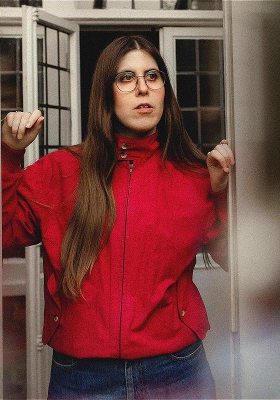 Ele stands in a doorframe with her arms up, leaning against it. They are wearing a red jacket. 