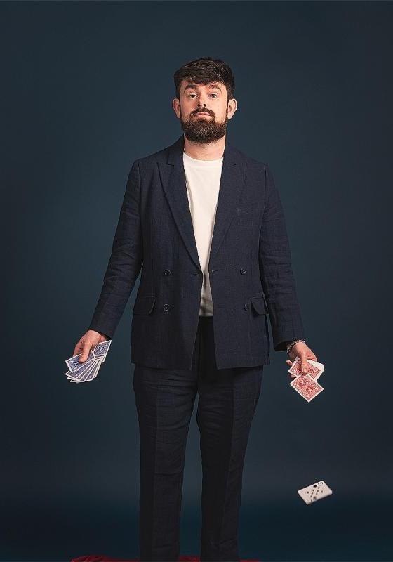 Tom in a black suit jacket, holding playing cards in both hands and one card is falling to the floor