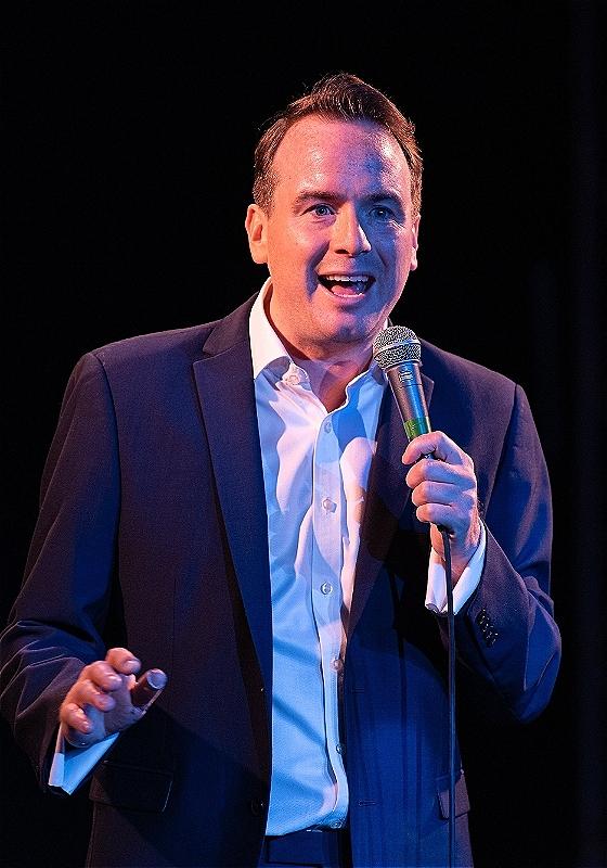 A stage picture of Matt Forde, a white man with brown hair wearing a white shirt and black suit jacket, speaking into a hand held mic, in front of a black background.