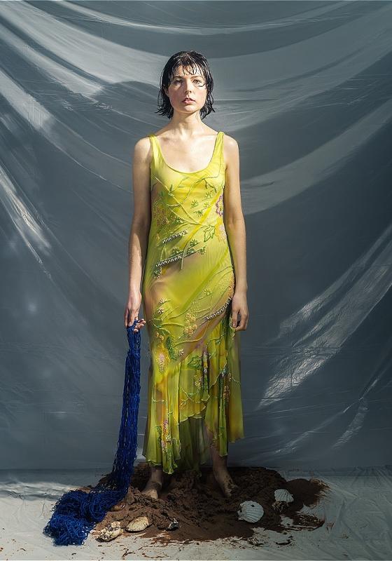 The performer stands in a sheer yellow/green dress and holding a blue fishing net trailing on the floor and standing in a small pile of mud.