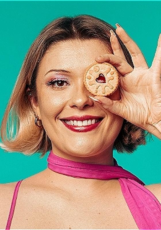 Cecily against a bright blue background, smiling and holding up a jammie dodger over one of her eyes. 
