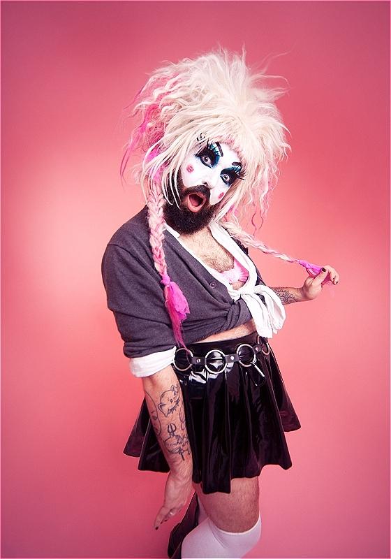 Baby Lame standing in front of a pink backdrop wearing Britney Spears' iconic schoolgirl outfit and a pink voluminous wig with pigtails. 