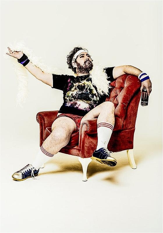Nick Helm, a white man with brown hair and a beard is sitting on a red padded leather chair with one leg on the armrest. He is wearing a white boa, sports cuffs and headband, sports socks and Converse shoes, and holding a Monster can in his left hand. He is looking up with his arms open.