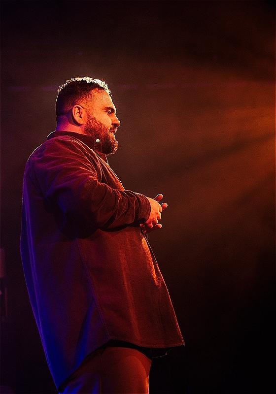 A stage picture of a brown man's profile, he has a short beard and a burgundy shirt, with a orange light in the background.