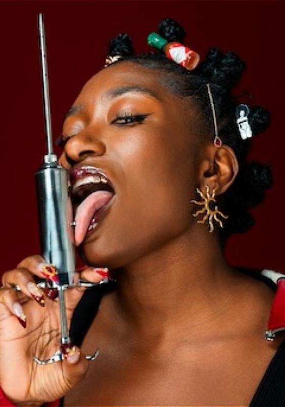 Sophie Duker, against a burgundy background, licking a large metal syringe that she is pointing upwards