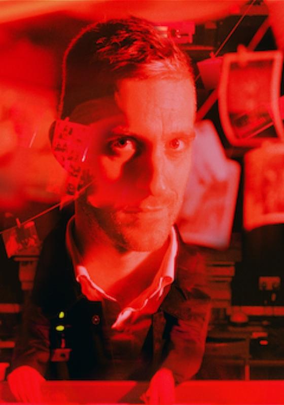 Joseph, bathed in red light, surrounded by hanging film photos.