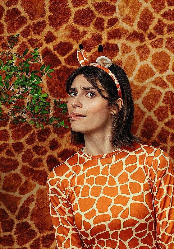 A woman wearing a giraffe patterned top and fake giraffe ears is licking a leaf on a branch that is coming into frame from the left. The background is giraffe pattern