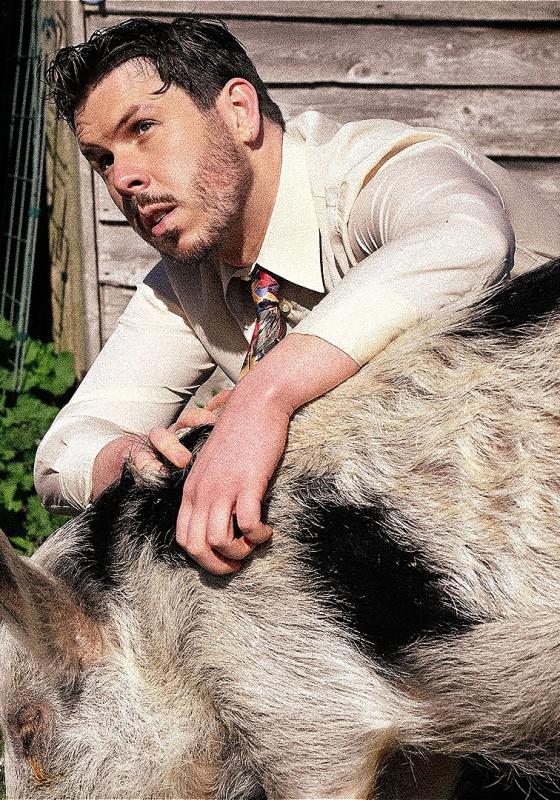 Dom crouched down behind a pig, wearing a white shirt and tie. 
