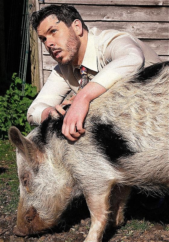 Dom crouched down behind a pig, wearing a white shirt and tie. 