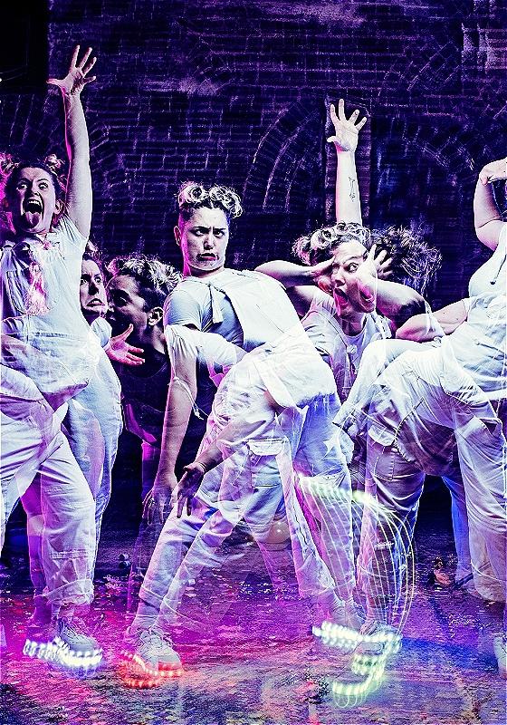 Four white performers dressed in white dungarees and t-shirts and wearing lit-up white shoes are dancing in different positions overlayed with each other. They stand in front of a dark purple brick wall.