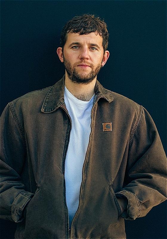 Rory leaning against a dark blue wall staring ahead into the camera wearing a Carhartt jacket.