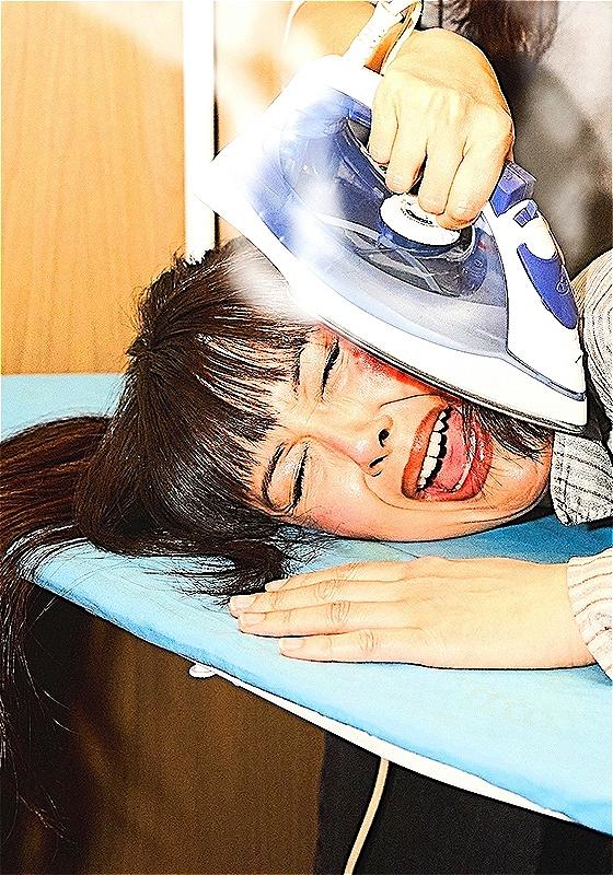 Patti Harrison's face is being pressed onto an ironing board by an unseen person, ironing her cheek with a blue and white iron. She has long brown hair and bangs and she is closing her eyes and opening her mouth as if she's screaming.