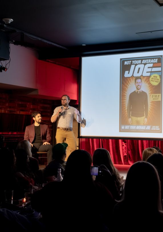 A man standing on stage holding a microphone whilst their friend sits beside them. A powerpoint slide is on the projector.
