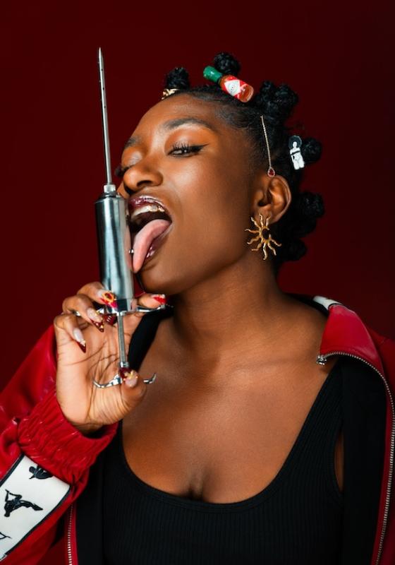 Sophie Duker, against a burgundy background, licking a large metal syringe that she is pointing upwards