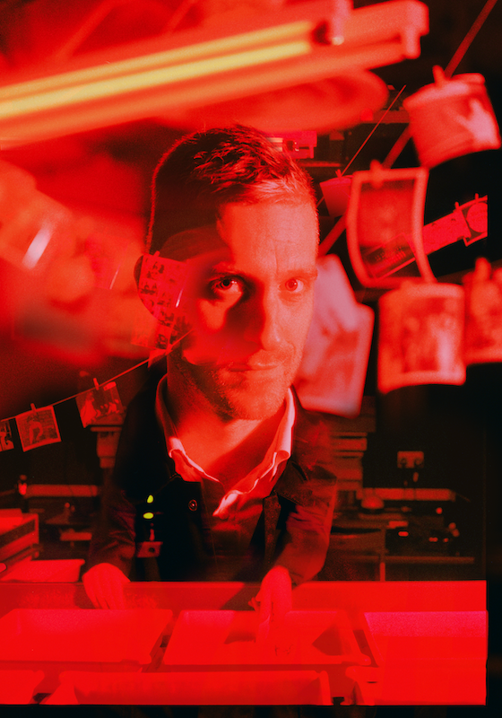 Joseph in a dark room with Polaroids developing with a red tint over the image