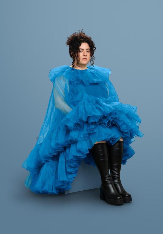 Jess wearing a blue tulle ruffled gown and a pair of leather knee-high boots sat on a block with with a vacant expression at the camera.