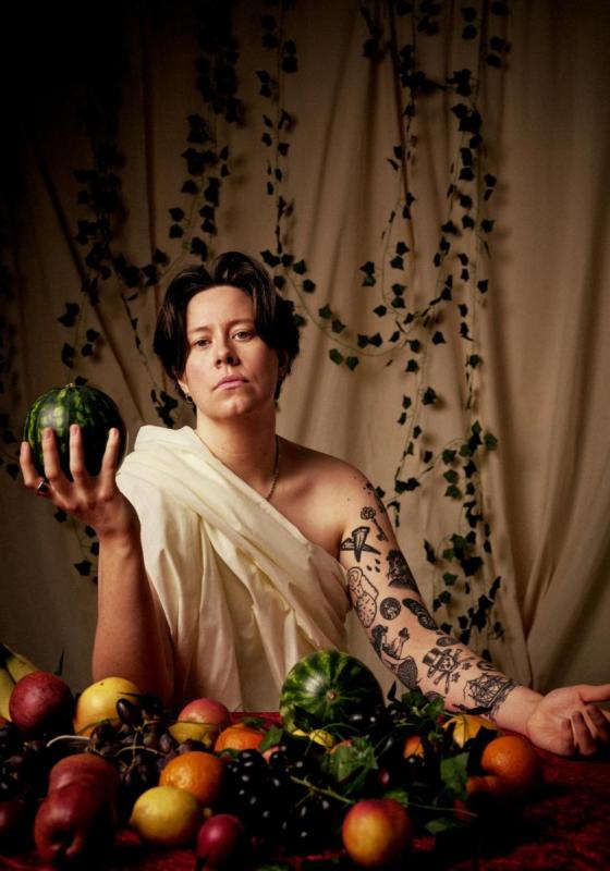 Chloe, holding a melon, wearing a toga sat in front of a table adorned with fruit. The curtain backdrop has leaves hanging in front. 