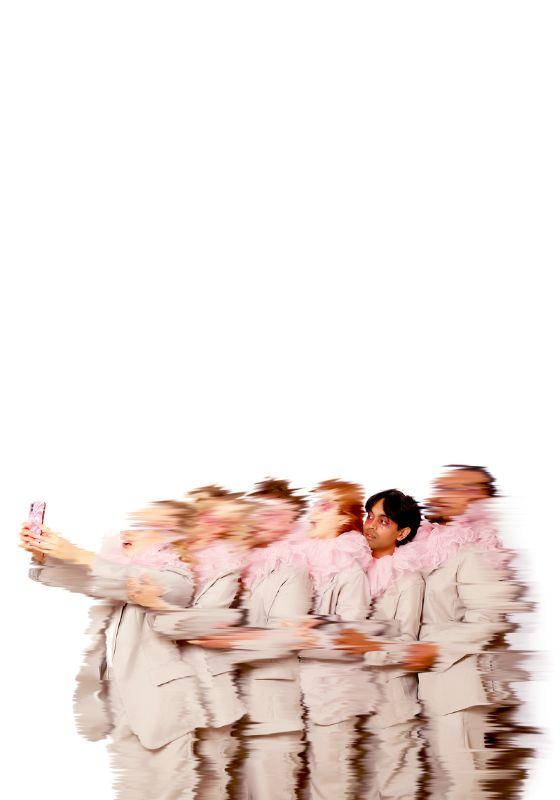 A blurry image of a line of performers standing one behind the other, wearing grey suits and pink ruffled collars. The person at the front is holding up a phone that they are all looking into 