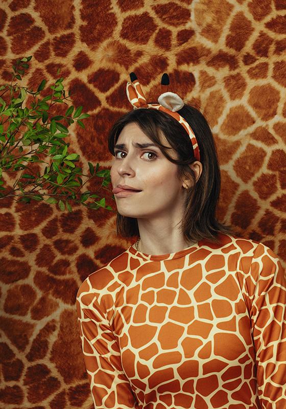 A woman wearing a giraffe patterned top and fake giraffe ears is licking a leaf on a branch that is coming into frame from the left. The background is giraffe pattern