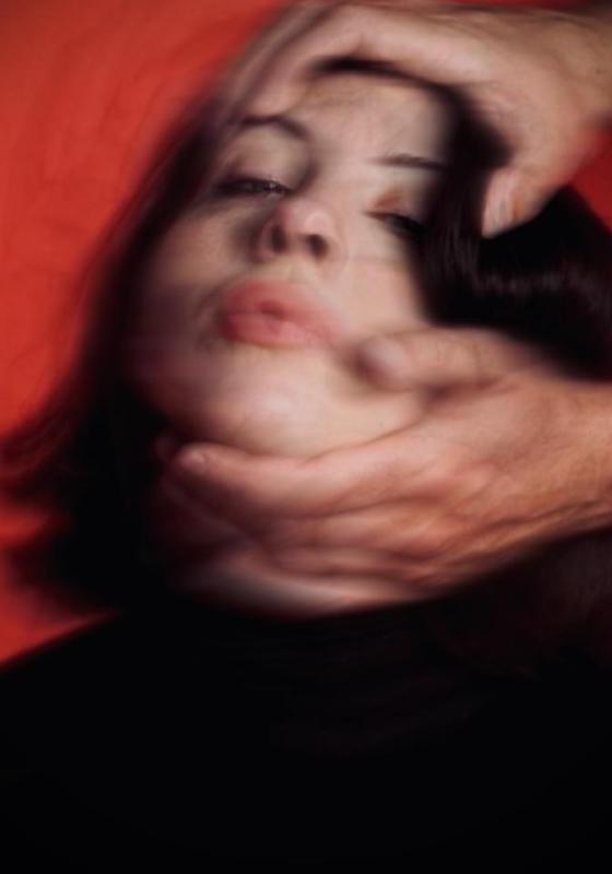 A blurry image of Harriet's face against a red background. Someone's hands reach in from out of frame and are holding the top of her head and her chin, pulling her head back so her face is tilted upwards