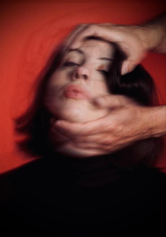 A blurry image of Harriet's face against a red background. Someone's hands reach in from out of frame and are holding the top of her head and her chin, pulling her head back so her face is tilted upwards
