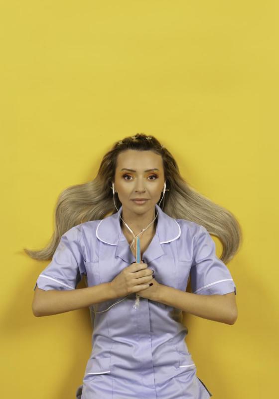 Kimberley is against a bright yellow background. They are wearing a blue nurses dress, with wired earphones in their ears. They are clasping a stick of rock between their hands, in front of their chest.