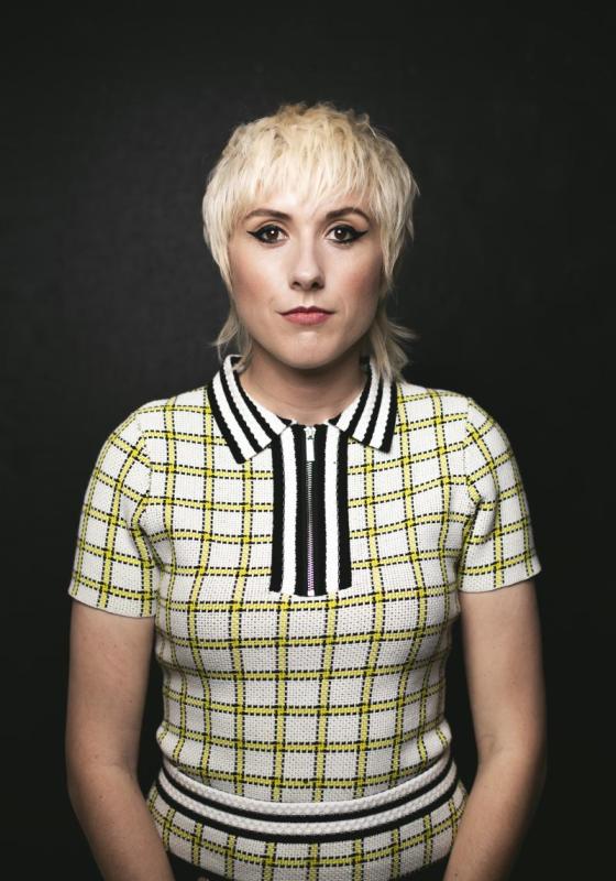 Maisie standing against a black background looking straight ahead. They are wearing a white t shirt with a yellow check pattern and black and white stripes on the collar