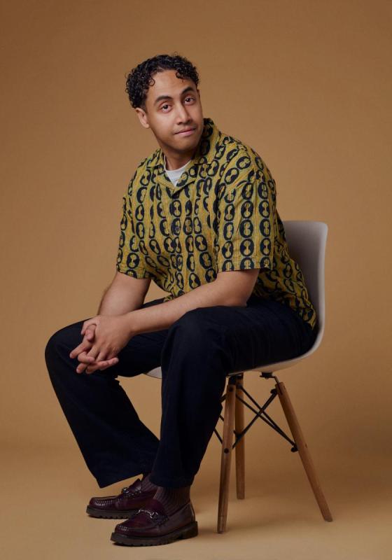 Omar sitting on a chair with his hands clasped gently in his lap. He wears a fun printed shirt against a beige brown background.