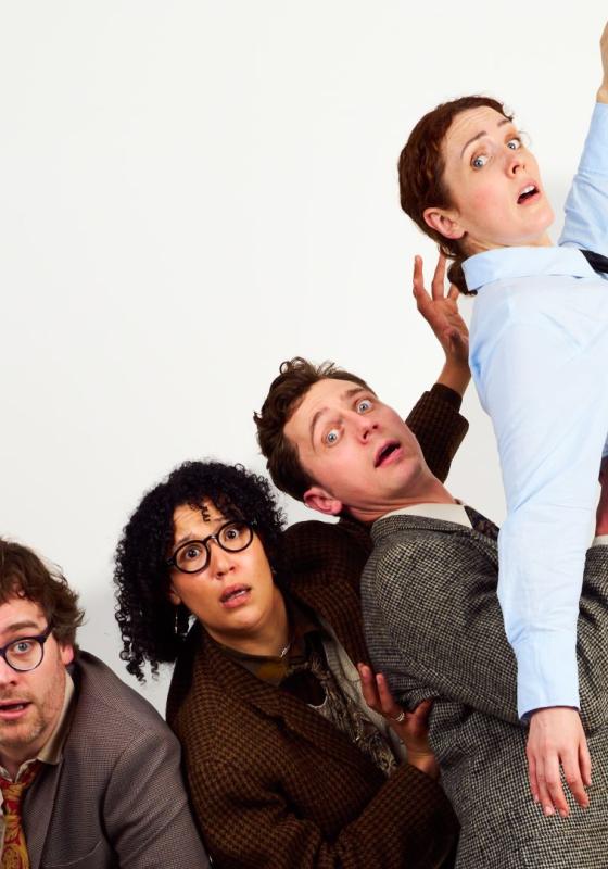 Cast of 4 performers dressed in suits and blazers looking alarmed against a plain white backdrop.