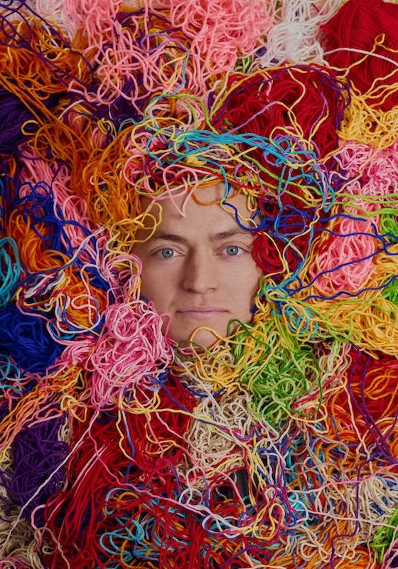 Tom's face is visible under a huge pile of different brightly coloured yarn 
