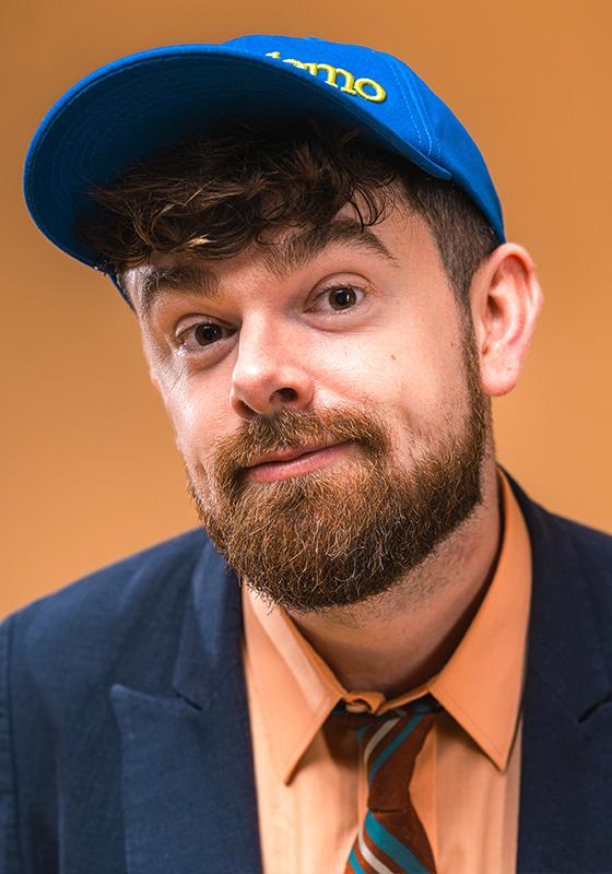 Tom brace dressed in a navy suit, orange shirt and tie and blue baseball cap on an orange background