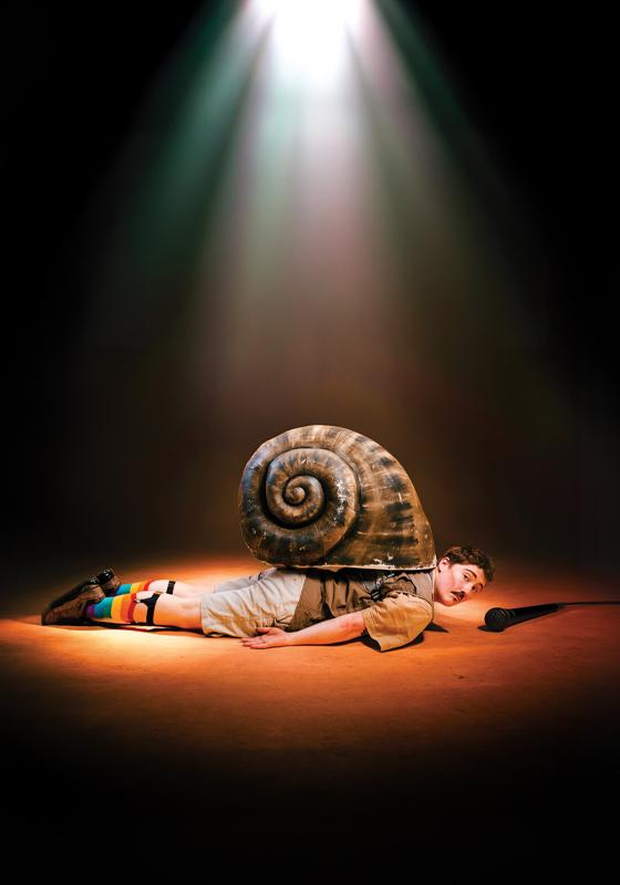 The performer lying flat with a large snail shell on their back. A microphone lies on the floor ahead of them.