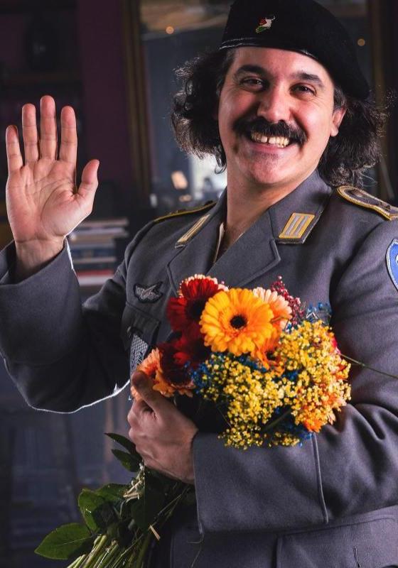Sami is wearing a military uniform, holding a bouquet of orange and red flowers. They are holding a hand up, waving to the viewer, and smiling. 
