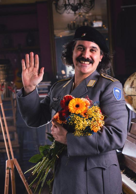 Sami is wearing a military uniform, holding a bouquet of orange and red flowers. They are holding a hand up, waving to the viewer, and smiling