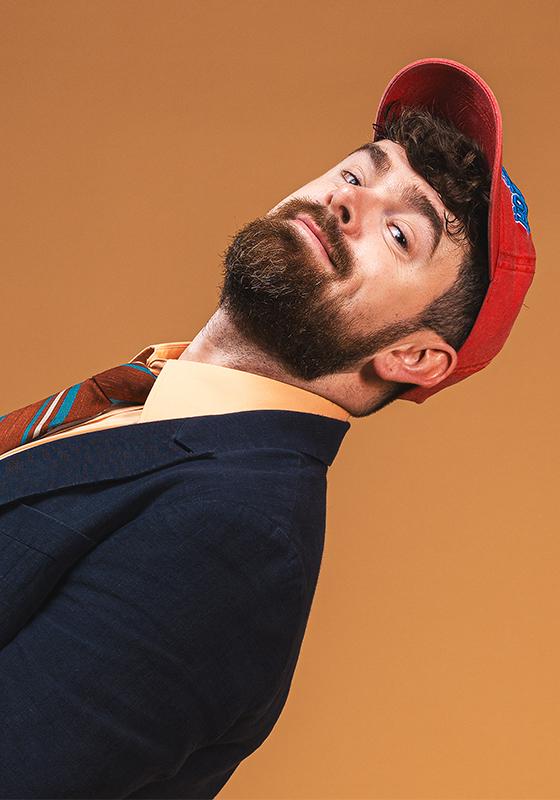 Tom brace dressed in a navy suit, orange shirt and tie and red baseball cap leaning back into frame agains an orange background
