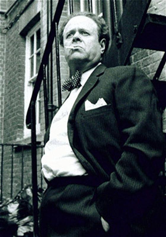A Black and white image of a smartly dressed man leant against a metal staircase as a cigarette dangles from his mouth.