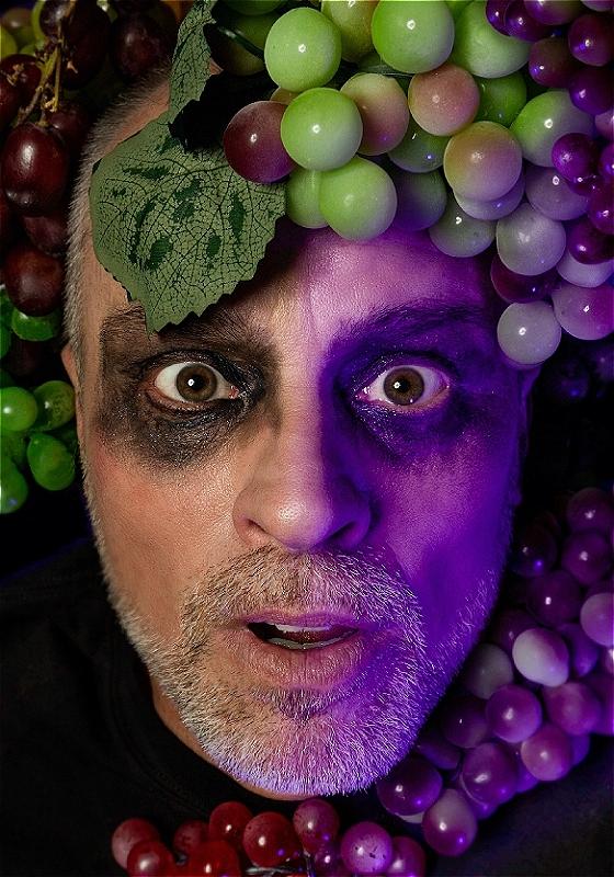 A man with a short white beard, brown eyes and black eyeshadow all around his eyes, is looking at the camera with bright eyes and his mouth slightly open. On his forehead and around his neck are white and red grapes and an ominous purple hue is lighting half of his face.
