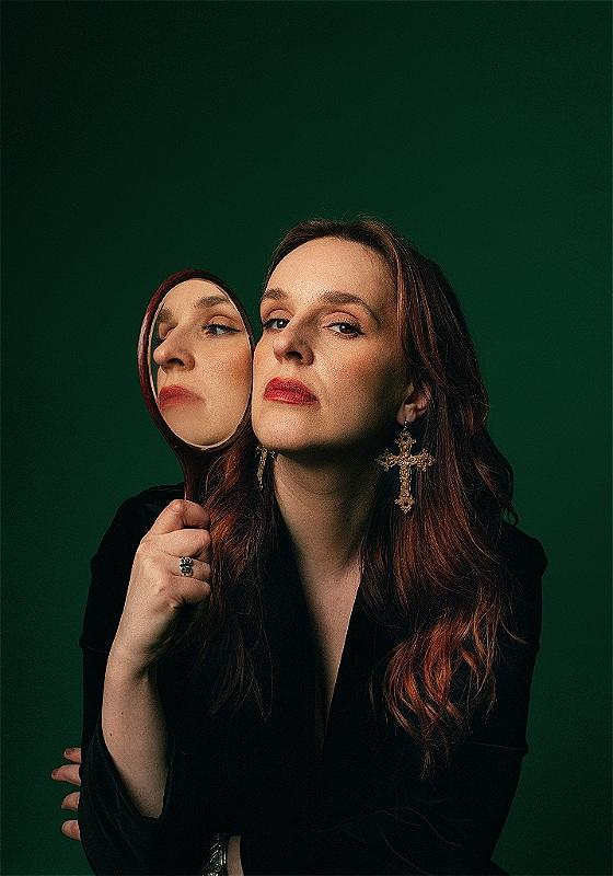 Katie Norris, a white woman with long brown hair, wearing red lipstick, gilded crucifix earrings and a black top, is looking at the camera holding a hand mirror to the side of her face so that the reflection of her face is looking to the side.