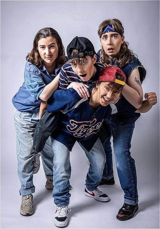 Four drag kings wearing 90s clothing stand huddled together posing like a stereotypical boyband. They look cheeky and youthful.  