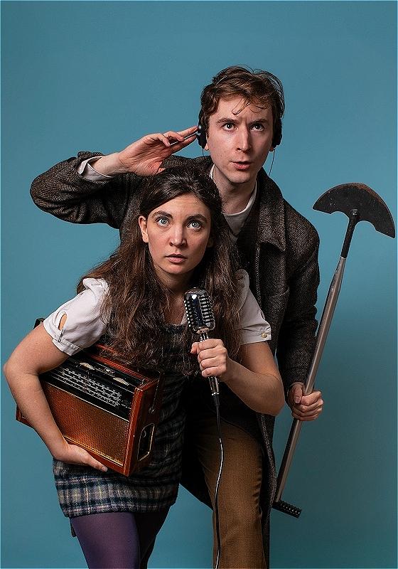 A white woman with long brown hair and a white man with short brown hair are standing together looking up intensely, in front of a blue background. The woman is holding an old timey radio and microphone, and the man is holding a shovel and wearing black headphones.