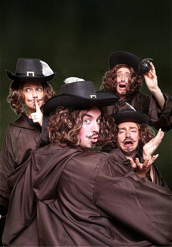 Four white performers all in Guy Fawkes drag, wearing black hats, long brown wigs and drawn goatees, are standing in various theatrical positions over a dark background.