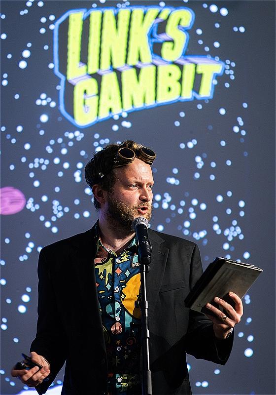 A man stands in front of a mic with a book in his hand and space goggles on his head, in front of a starry background. 