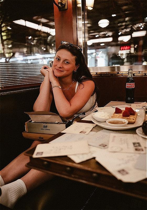 A woman sat in a warmly lit restaurant smiles at the camera. She is sat on a table with a typewriter on her lap. On the table there is a plate of food, a glass bottle of coca cola and a collection of letters.